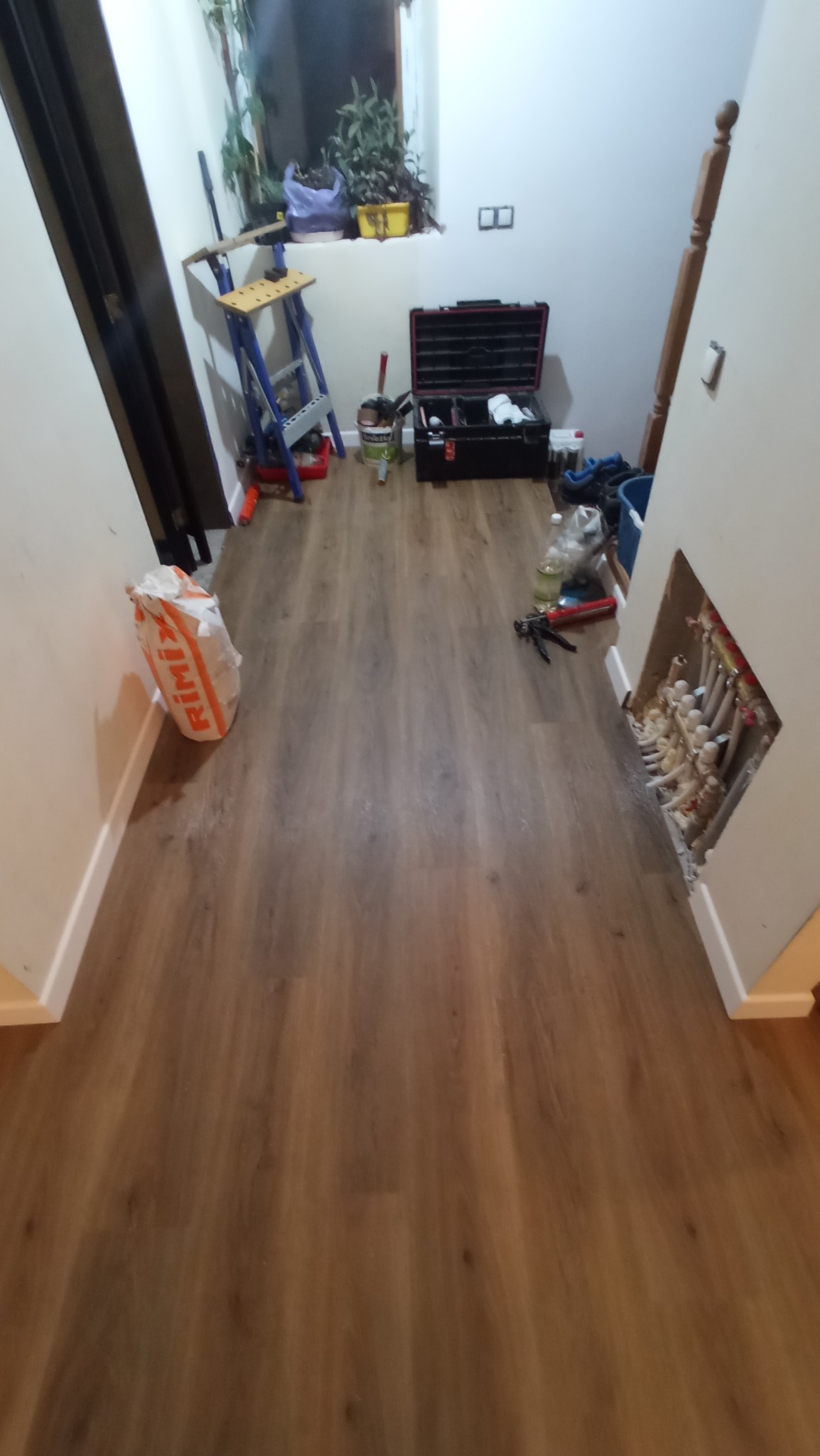 Ułożone panele podłogowe w korytarzu, z widoczną torbą z klejem, skrzynką z narzędziami, drabinką i instalacją grzewczą w ścianie.