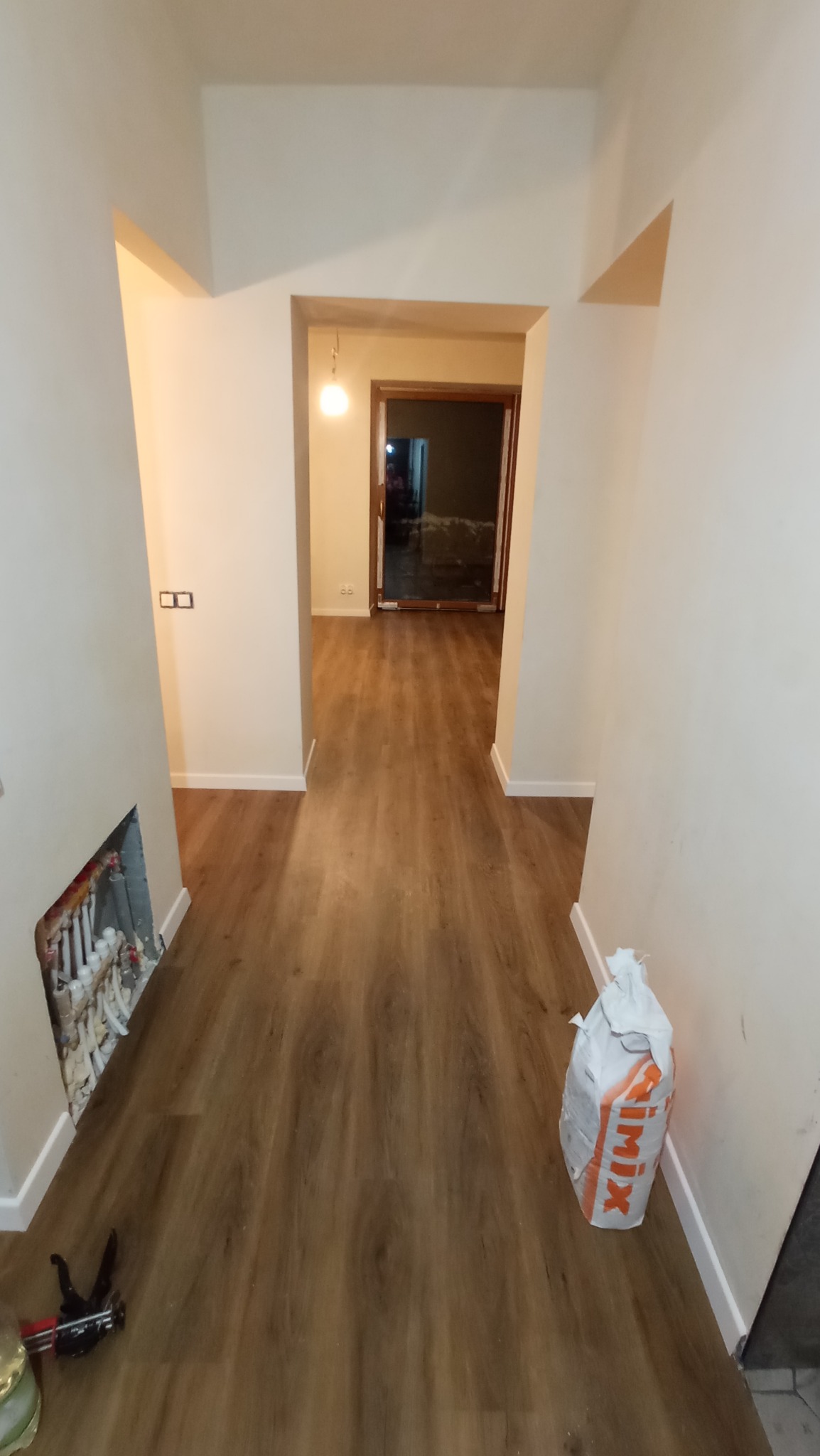 Korytarz z nowo ułożonymi panelami podłogowymi w odcieniach brązu, widoczny worek zaprawy SIMIX i narzędzia przy ścianie oraz niedokończone prace przy instalacji grzewczej w ścianie.