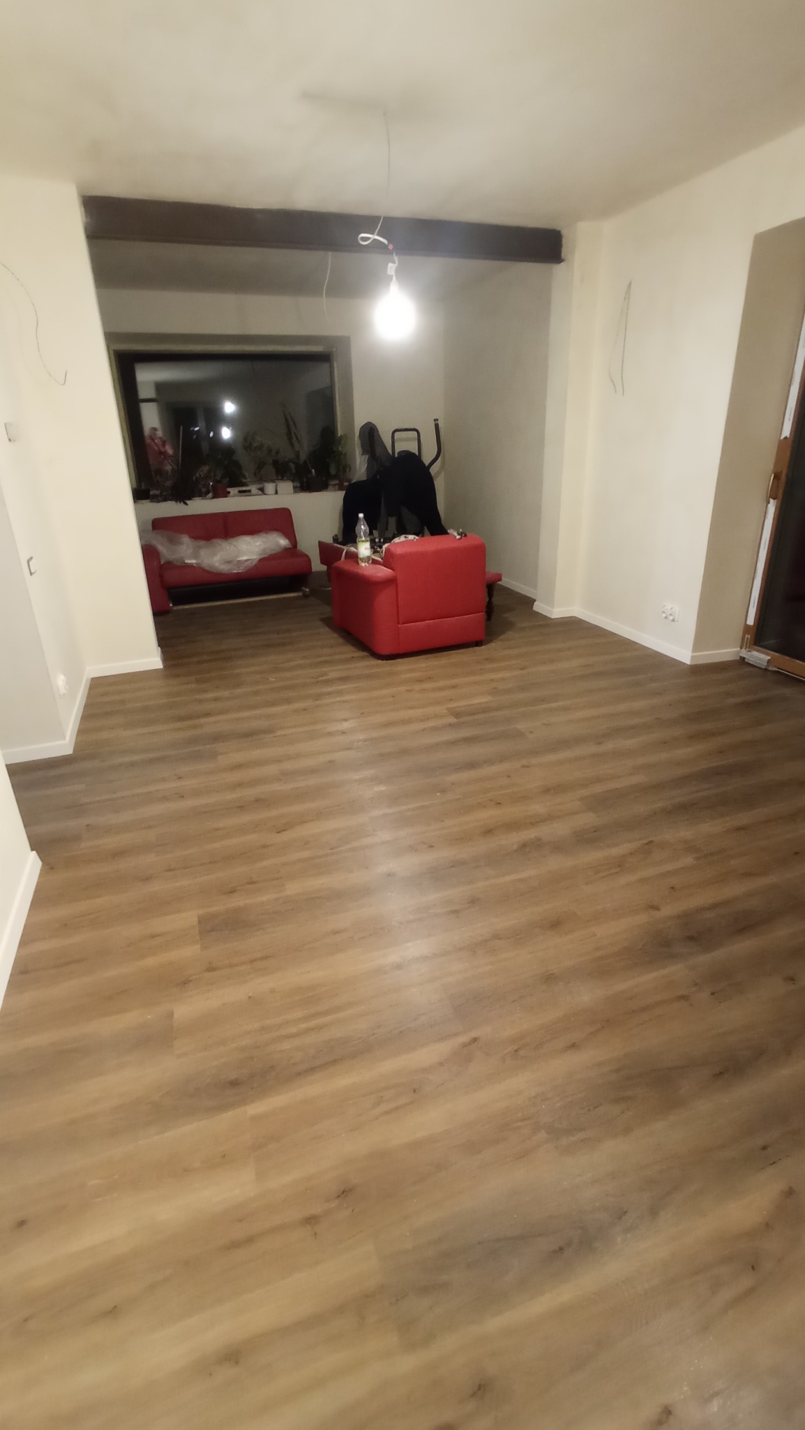 Jasna podłoga z paneli drewnopodobnych w pokoju z czerwonymi meblami, widok na sofę przykrytą folią ochronną i sprzęt do ćwiczeń w tle.