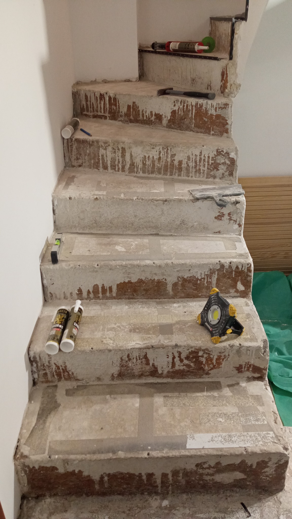 Schody betonowe w trakcie remontu, z widocznymi śladami po starych płytkach i pozostałościami kleju, narzędzia pracy: poziomica, lampa robocza, młotek, szpachelka, tuby z klejem lub silikonem.