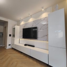 Piotr Kaczor-Usługi Remontowo Budowlane - Nowoczesny salon z białą zabudową meblową, marmurową ścianą TV i parkietem w jodełkę. Oświetlenie punktowe podkreśla dekoracyjne panele ścienne.
