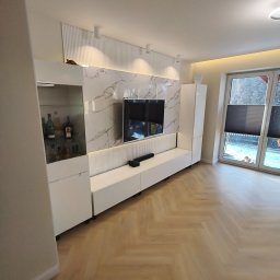 Piotr Kaczor-Usługi Remontowo Budowlane - Nowoczesny salon z białą zabudową meblową pod telewizor, marmurowym panelem ściennym i parkietem w jodełkę. Widoczne oświetlenie LED i dekoracje.