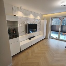 Piotr Kaczor-Usługi Remontowo Budowlane - Nowoczesna aranżacja salonu z białą zabudową meblową, marmurowym panelem ściennym i parkietem w jodełkę. Widoczne oświetlenie LED i dekoracyjne elementy.