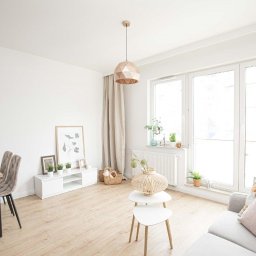 Piotr Kaczor-Usługi Remontowo Budowlane - Jasne, przestronne wnętrze salonu z drewnianą podłogą, geometrycznymi lampami wiszącymi, białymi meblami i dużym oknem z widokiem na zewnątrz, uzupełnione dekoracjami roślinnymi i tekstyliami...