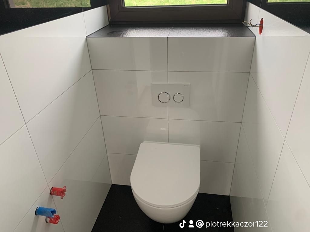 Podwieszana toaleta z białymi płytkami i czarną podłogą, widoczne wystające rury instalacyjne oraz niedokończone podłączenie elektryczne.