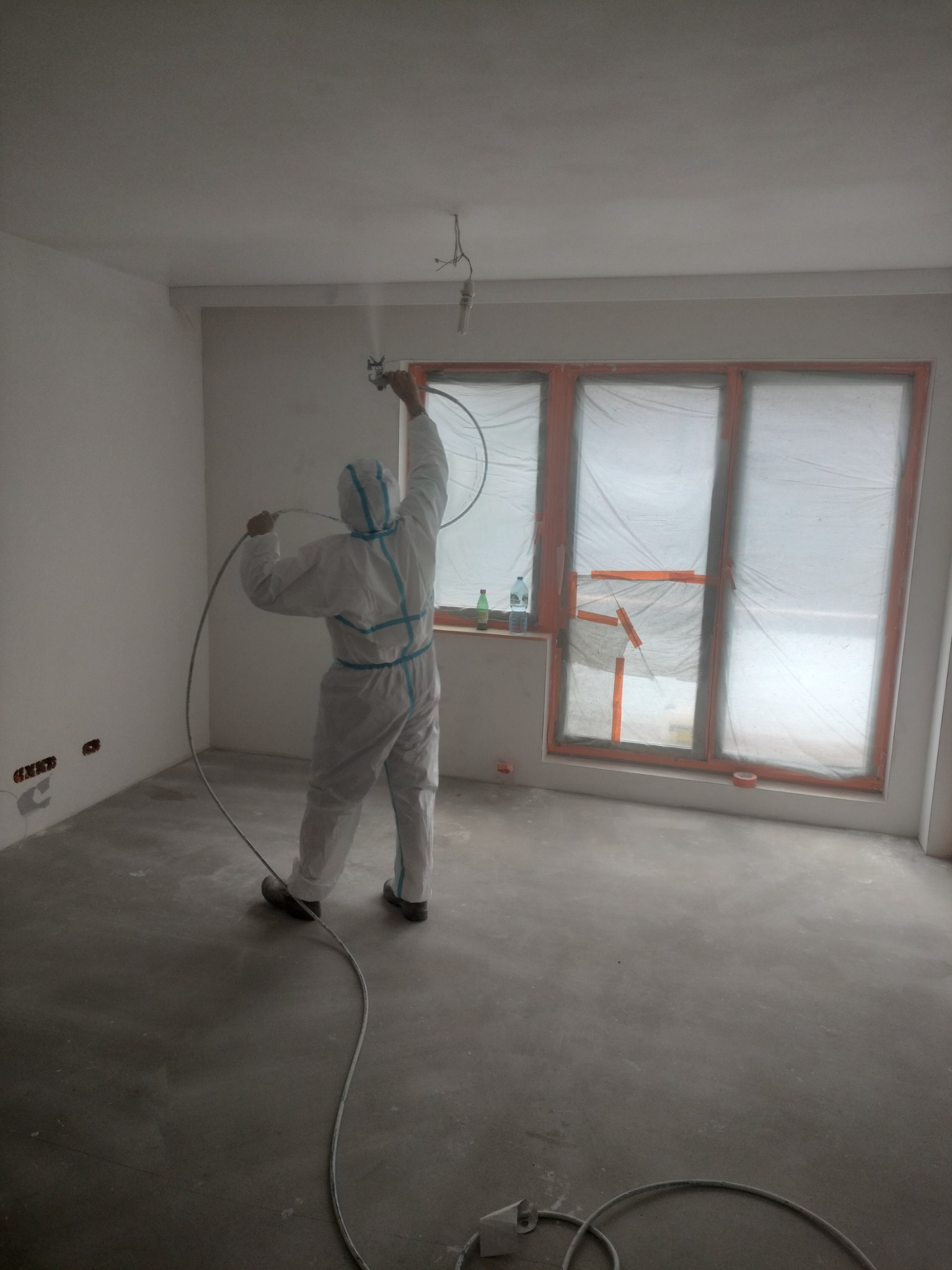 Pracownik w kombinezonie ochronnym nanosi natryskowo masę na ściany w pomieszczeniu z oknami zabezpieczonymi folią, widoczne kable i wąż od urządzenia.