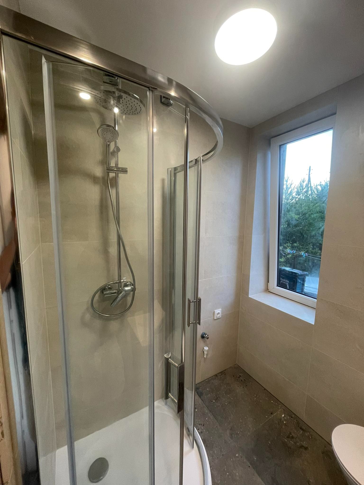 Nowoczesna kabina prysznicowa z zaokrąglonym szkłem, chromowanymi detalami i deszczownicą w wykończonej łazience z beżowymi płytkami i oknem z widokiem na zieleń.