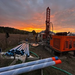 Techdrill Studnie głębinowe oraz pompy ciepła - Pomarańczowa wiertnica na gąsienicach na tle zachodzącego słońca, obok sterta niebieskich rur, pracownik w oddali przy wykopie.