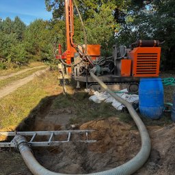 Techdrill Studnie głębinowe oraz pompy ciepła - Pomarańczowa wiertnica na gąsienicach przy wykopie, widoczna drabina w wykopie i wąż odprowadzający wodę, las w tle.