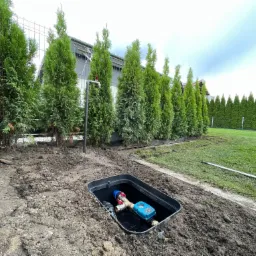 Instalacja wodomierza w czarnej skrzynce w ziemi, widoczny kran i rząd tuj w tle, częściowo zrekultywowany teren.