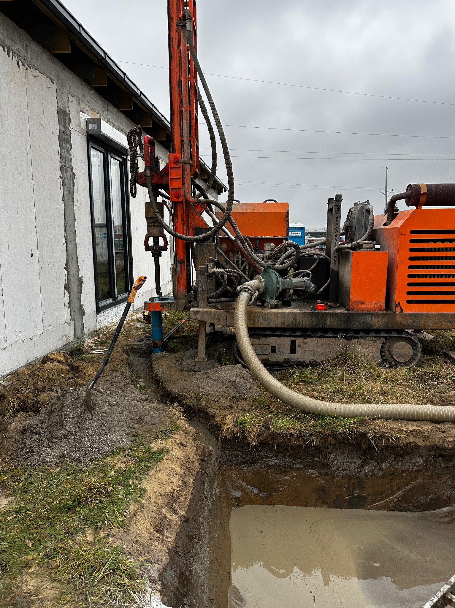 Pomarańczowa wiertnica obok budynku z wykopem wypełnionym wodą, łopata oparta o rurę, pochmurne niebo.