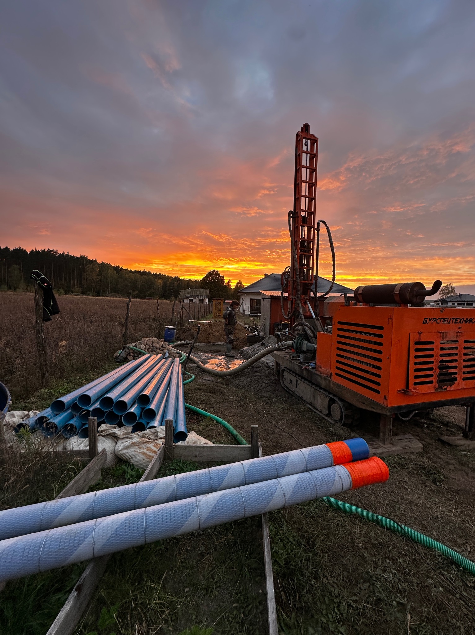 Pomarańczowa wiertnica na gąsienicach na tle zachodzącego słońca, obok sterta niebieskich rur, pracownik w oddali przy wykopie.