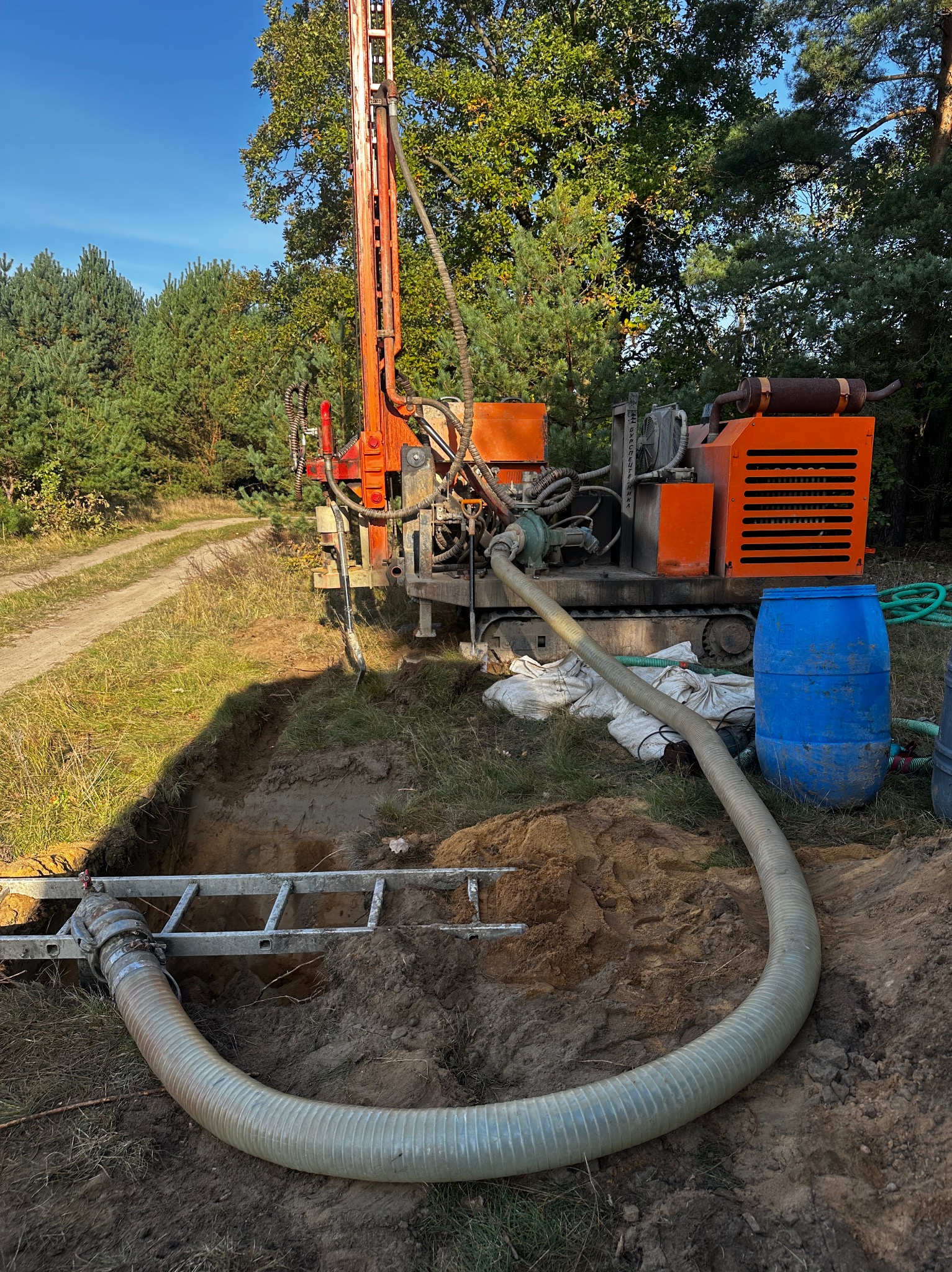 Pomarańczowa wiertnica na gąsienicach przy wykopie, widoczna drabina w wykopie i wąż odprowadzający wodę, las w tle.