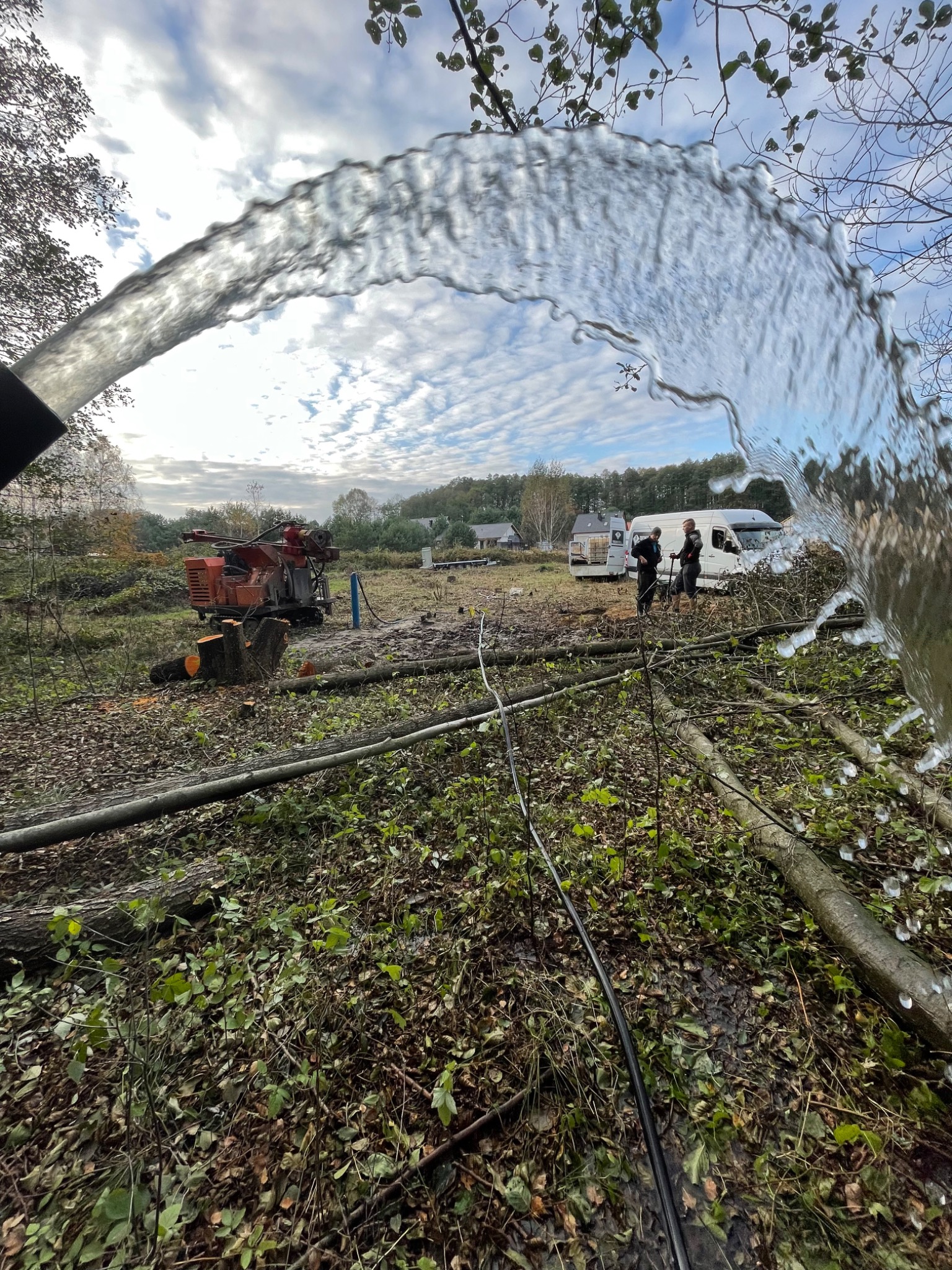 Strumień wody z węża podczas wiercenia, w tle pomarańczowa wiertnica, ekipa i samochody na wykarczowanym terenie.