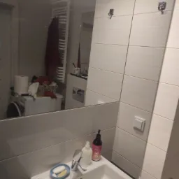 Umywalka w łazience z mydłem i płynem do dezynfekcji, w lustrze odbija się pralka z koszem pełnym prania oraz suszarka na ręczniki.