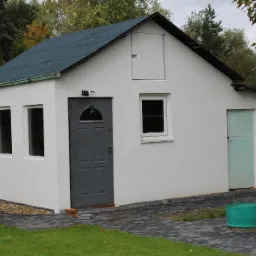 Biały budynek gospodarczy z szarymi drzwiami i niebieskim dachem, widoczne świeżo otynkowane ściany zewnętrzne i otoczenie z kostki brukowej.