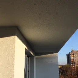 Balkon po remoncie z nową, jasną elewacją i świeżo pomalowanym sufitem, widok na budynek mieszkalny i drzewa jesienią.