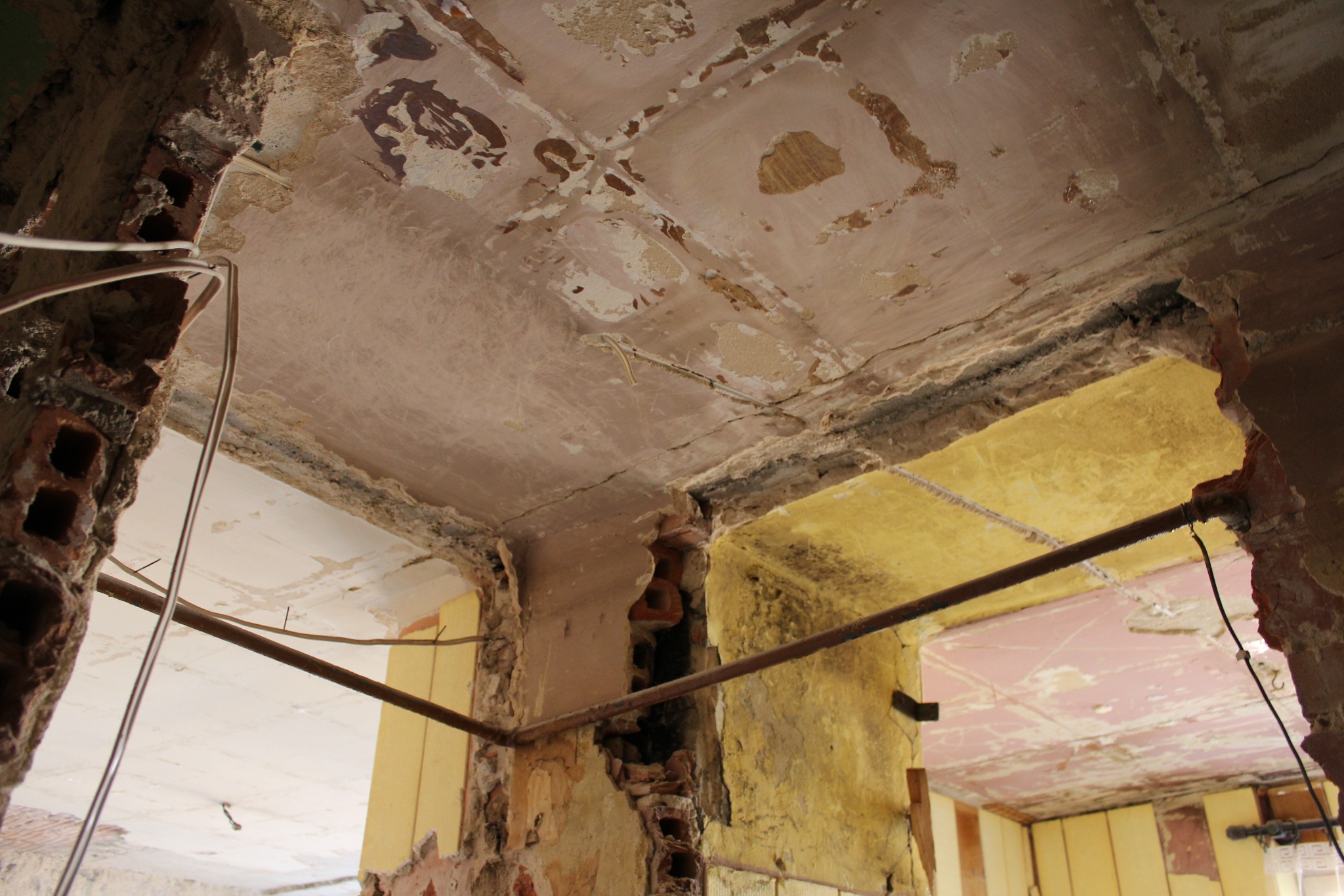 Fragment ściany i sufitu w trakcie prac remontowych, odsłonięta cegła, stara instalacja elektryczna, widoczne warstwy farby i tynku.