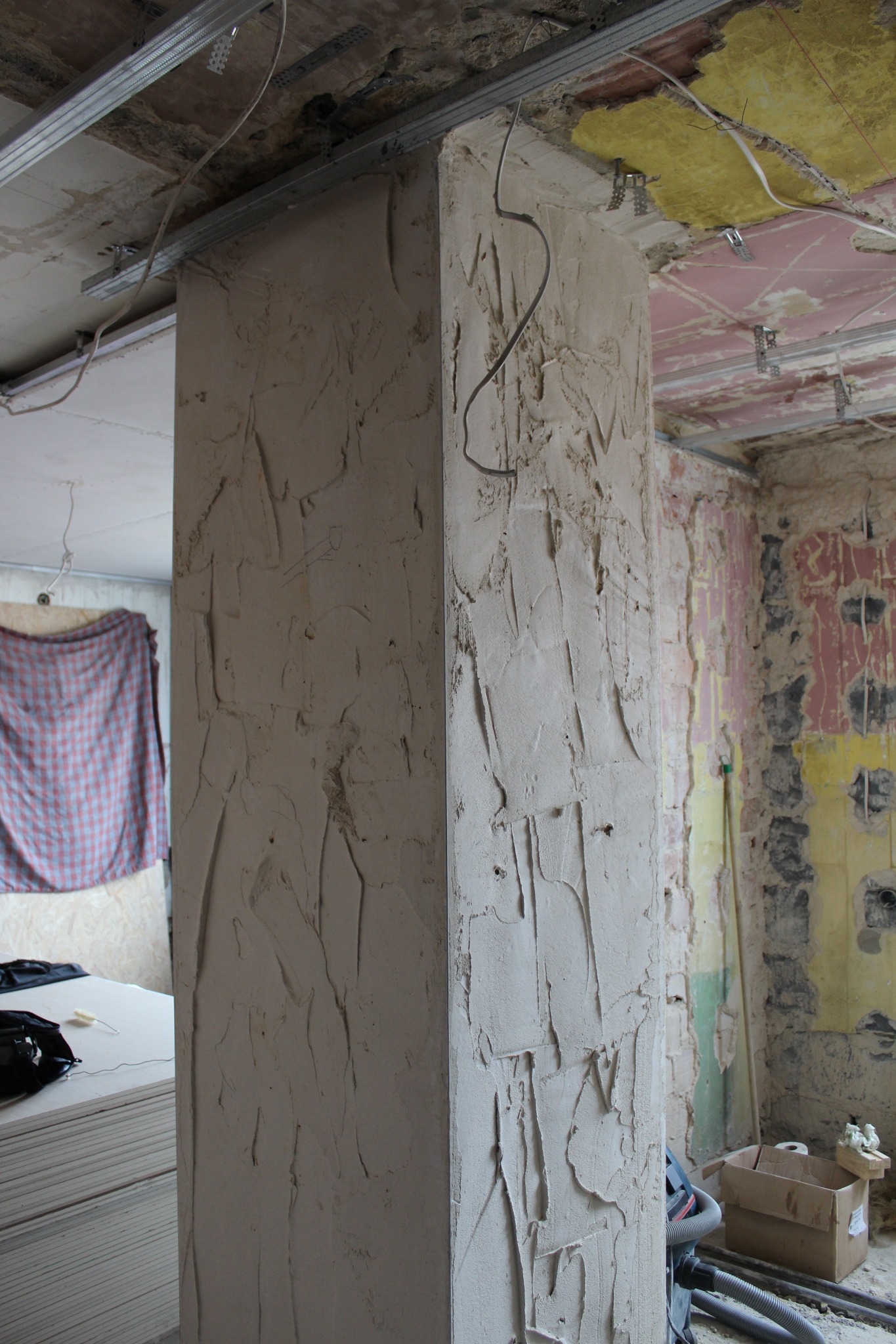 Narożnik filaru w trakcie tynkowania, widoczne surowe wykończenie, instalacje pod sufitem i częściowo zerwana tapeta w tle.