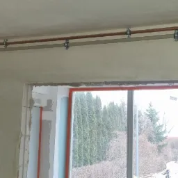 Montaż instalacji grzewczej w surowym stanie w Olsztynie, widoczne rury miedziane i plastikowe przymocowane do ściany nad oknem.