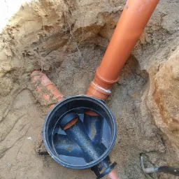 Studzienka kanalizacyjna z rurami z tworzywa sztucznego w wykopie ziemnym, widoczne ślady świeżego montażu i narzędzie.