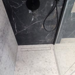 Adam Wieczorek - Nowoczesny prysznic z czarnymi marmurowymi płytkami na ścianach i jasną, wzorzystą podłogą, widoczny odpływ liniowy i fragment kabiny prysznicowej, dół kadru zajmują stopy w klapkach.