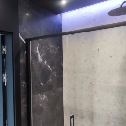 Adam Wieczorek - Nowoczesna kabina prysznicowa z czarnymi elementami, marmurowa ściana podświetlona od góry, szklane drzwi z minimalistycznym uchwytem.