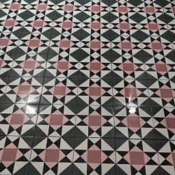 Adam Wieczorek - Wzorzysta podłoga z płytek w odcieniach różu, czerni i bieli, ułożona w geometryczny deseń, widoczna z perspektywy podłogi, z fragmentem szafki w tle i niebieskim wiaderkiem w rogu kadru.