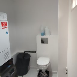 Adam Wieczorek - Biała toaleta wisząca z widocznymi rurami w czarnym pojemniku, obok pralka i suszarka w małej, białej łazience z jednym oknem.