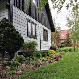 Adam Wieczorek - Jasnoszara drewniana elewacja domu z ciemnymi drewnianymi detalami, brązowe okna, widok na ogród z zielonym trawnikiem i rabatą kwiatową.