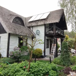 Dwukondygnacyjny dom z białymi ścianami, ciemnymi elementami drewnianymi i gontowym dachem, otoczony zielenią. Na dachu panele solarne. Przystawiona drabina sugeruje prace na elewacji.