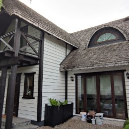 Adam Wieczorek - Biały dom z drewnianą elewacją i dachem krytym gontem, z ciemnobrązowymi wykończeniami, widok z zewnątrz, z widocznymi puszkami po farbie na ziemi przed domem, sugerującymi świeżo zakończone prace.