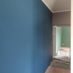 Adam Wieczorek - Świeżo pomalowana na niebiesko ściana w korytarzu z widoczną podłogą, zabezpieczoną folią malarską w tle, prowadząca do pokoju pomalowanego na zielono.