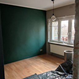 Adam Wieczorek - Pokój w trakcie remontu: ściana pomalowana na ciemnozielono, podłoga zabezpieczona folią ochronną, widoczna lampa wisząca i okno z grzejnikiem.