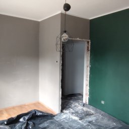 Adam Wieczorek - Pokój w trakcie malowania: jedna ściana szara, druga zielona, zabezpieczona podłoga, widoczna surowa ościeżnica i nowoczesna lampa.