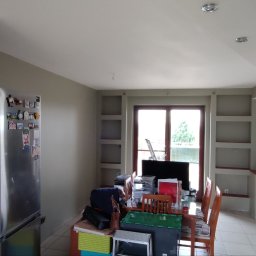 Adam Wieczorek - Wnętrze w trakcie remontu: ściany w odcieniach szarości, zabudowane półki, stół z krzesłami częściowo zastawiony sprzętem i materiałami budowlanymi, widoczna lodówka z magnesami i palnik na półce.