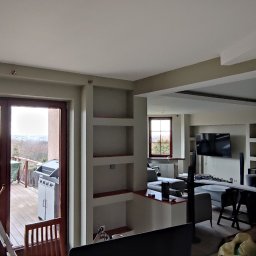 Adam Wieczorek - Wnętrze mieszkania po odświeżeniu ścian, widoczne wnęki w ścianach, drabina przy wyjściu na balkon i częściowo umeblowany salon w głębi.