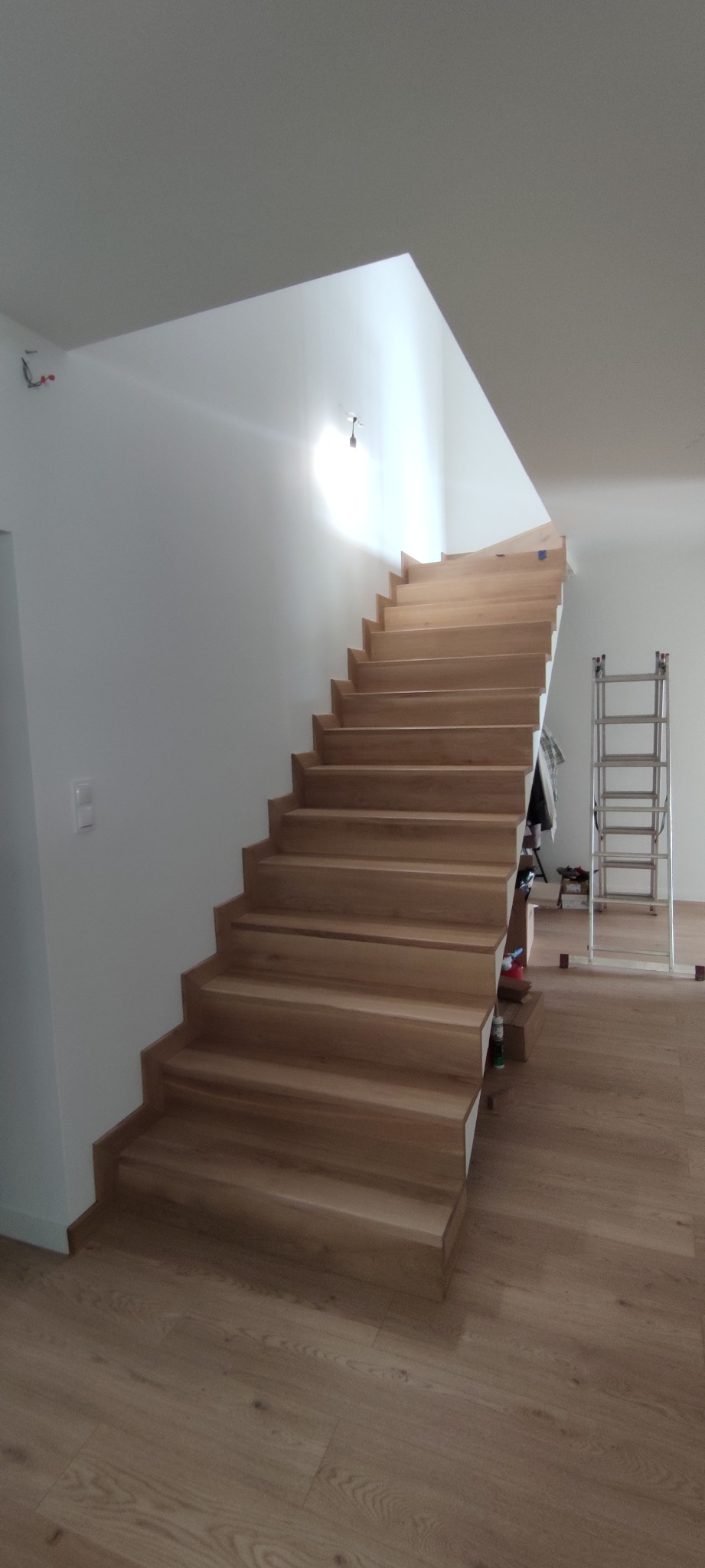 Jasne, drewniane schody w nowoczesnym wnętrzu, widoczne z dołu, z białą ścianą i podłogą z paneli, drabina aluminiowa w tle.