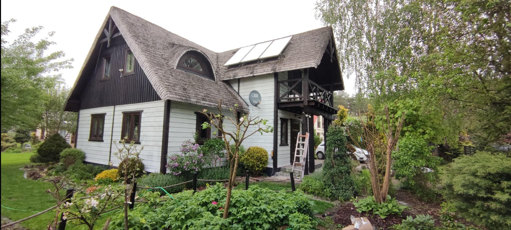 Dwukondygnacyjny dom z białymi ścianami, ciemnymi elementami drewnianymi i gontowym dachem, otoczony zielenią. Na dachu panele solarne. Przystawiona drabina sugeruje prace na elewacji.