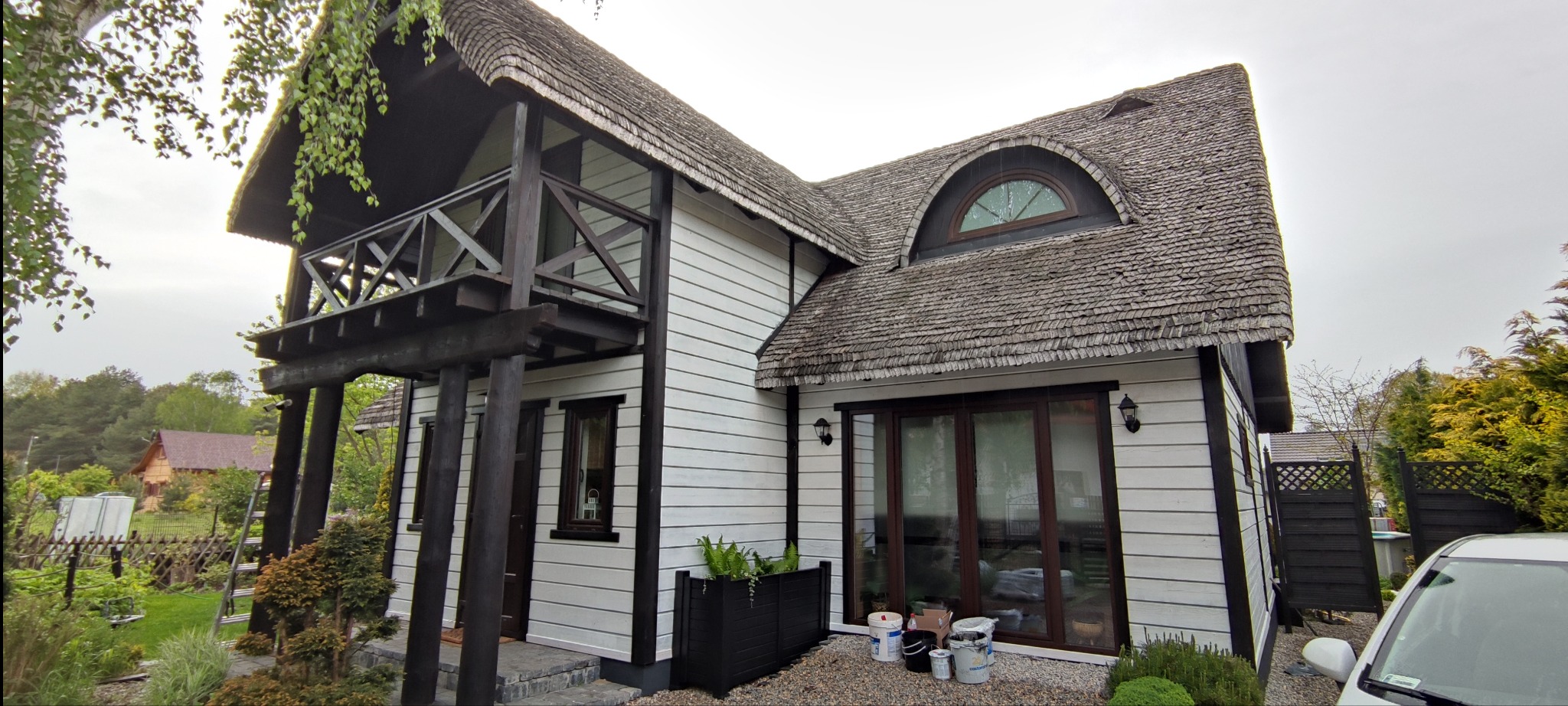 Biały dom z drewnianą elewacją i dachem krytym gontem, z ciemnobrązowymi wykończeniami, widok z zewnątrz, z widocznymi puszkami po farbie na ziemi przed domem, sugerującymi świeżo zakończone prace.