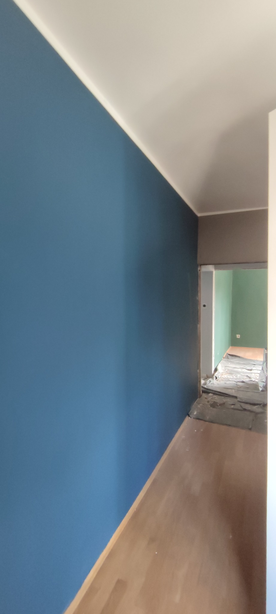 Świeżo pomalowana na niebiesko ściana w korytarzu z widoczną podłogą, zabezpieczoną folią malarską w tle, prowadząca do pokoju pomalowanego na zielono.