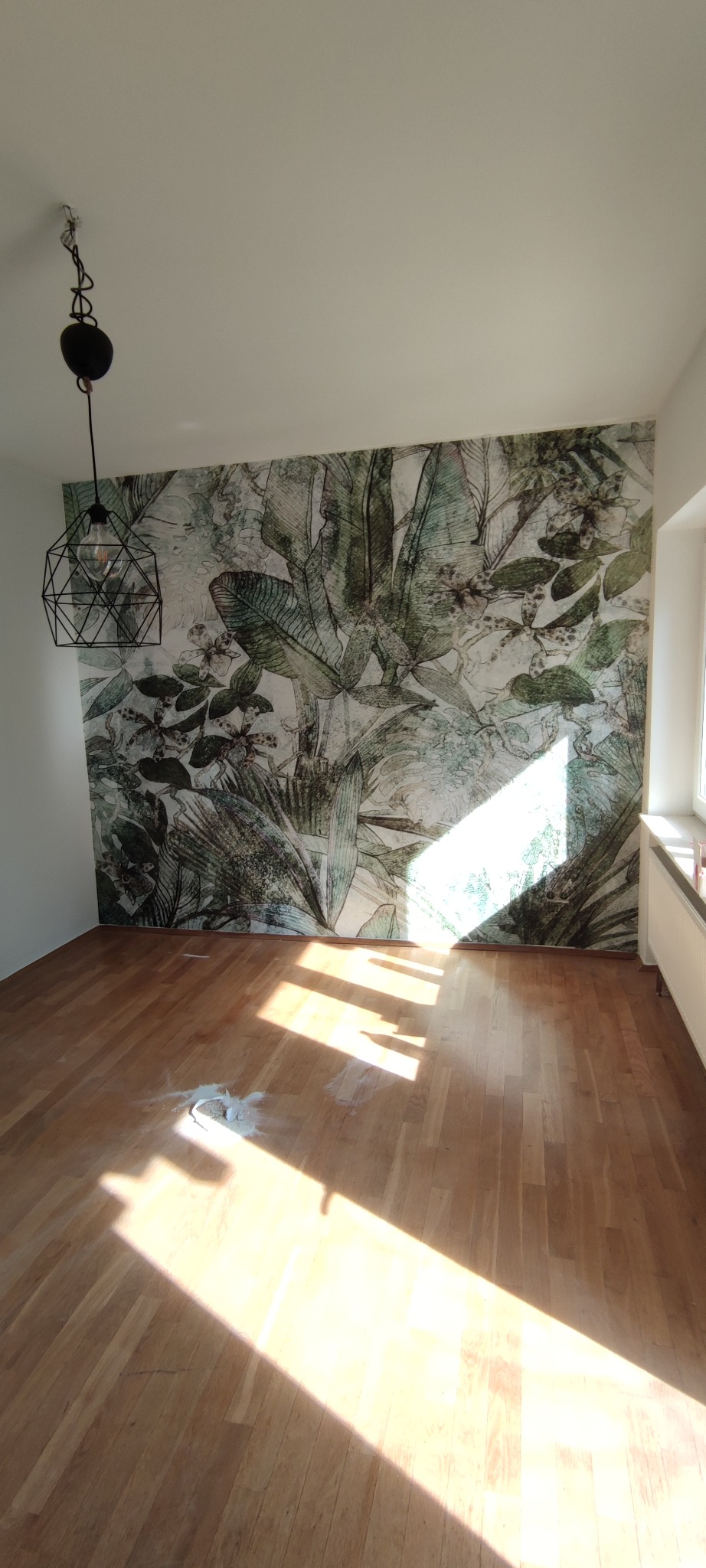 Pusty pokój z drewnianą podłogą, na ścianie fototapeta z motywem tropikalnych liści, wisząca lampa w stylu industrialnym i plama gipsu na podłodze, oświetlony silnym światłem słonecznym.