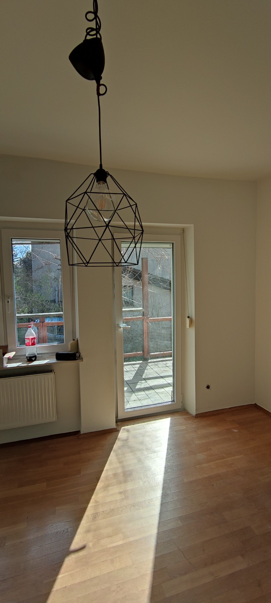 Jasne wnętrze pokoju z parkietem, oświetlone popołudniowym słońcem wpadającym przez okno balkonowe, na parapecie butelka Coca-Coli, a z sufitu zwisa nowoczesna lampa z odsłoniętą żarówką.