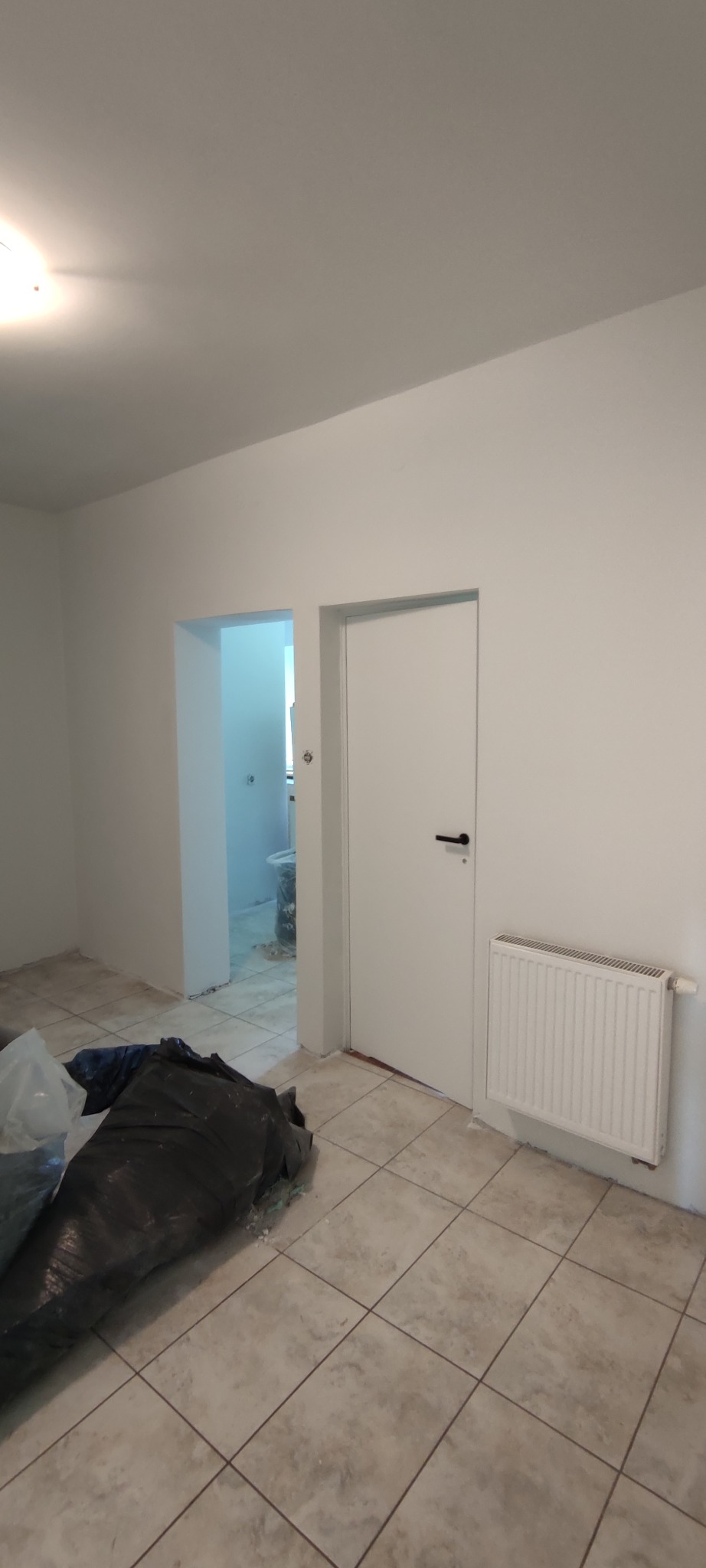 Wnętrze mieszkania po malowaniu ścian na biało, widoczny grzejnik, drzwi i podłoga wyłożona płytkami, zabezpieczona folią
