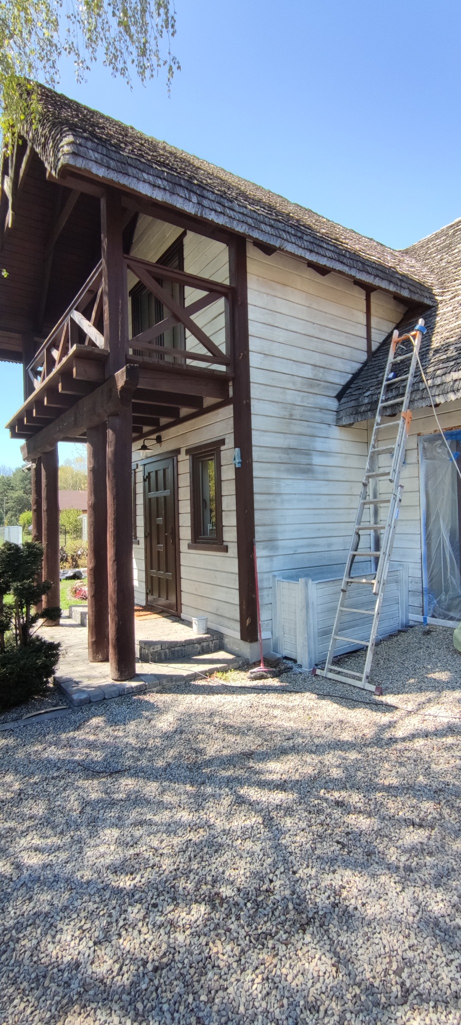 Dom z drewnianą elewacją w trakcie malowania na jasny kolor, widoczna drabina oparta o ścianę, fragmenty elewacji jeszcze niepomalowane, ukazujące pierwotny odcień drewna.