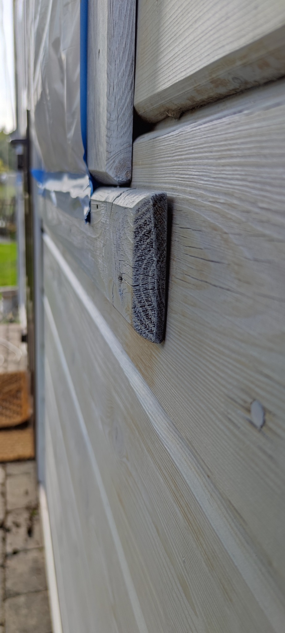 Fragment drewnianej elewacji domu, częściowo zabezpieczonej folią i taśmą malarską, w trakcie malowania na jasny kolor.