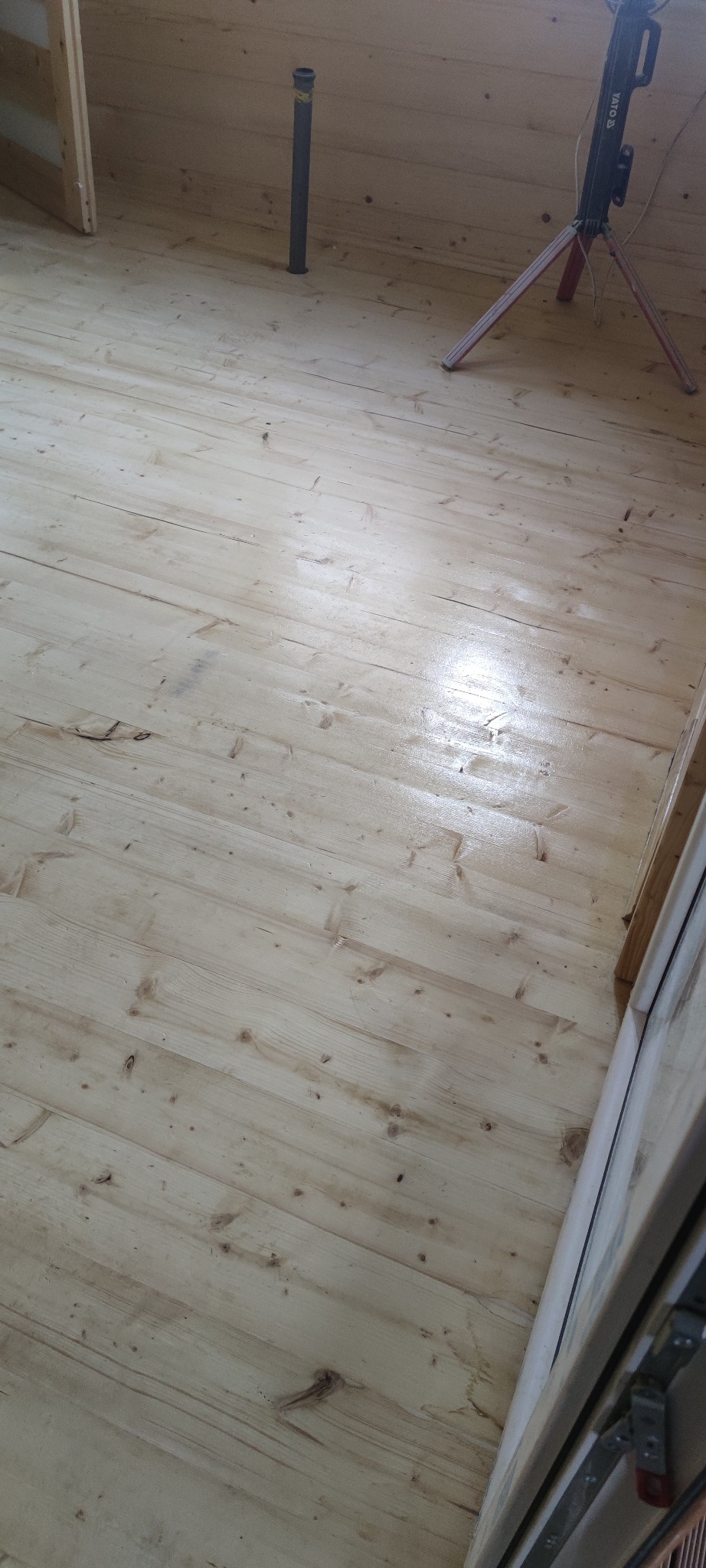 Jasne, świeżo położone panele podłogowe z widocznym oświetleniem budowlanym i fragmentem rury wentylacyjnej w tle.