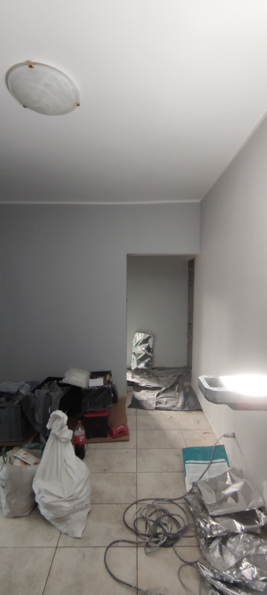 Szare ściany w trakcie odświeżania, zabezpieczona folią podłoga, narzędzia i materiały budowlane w nieładzie, widoczna lampa sufitowa.