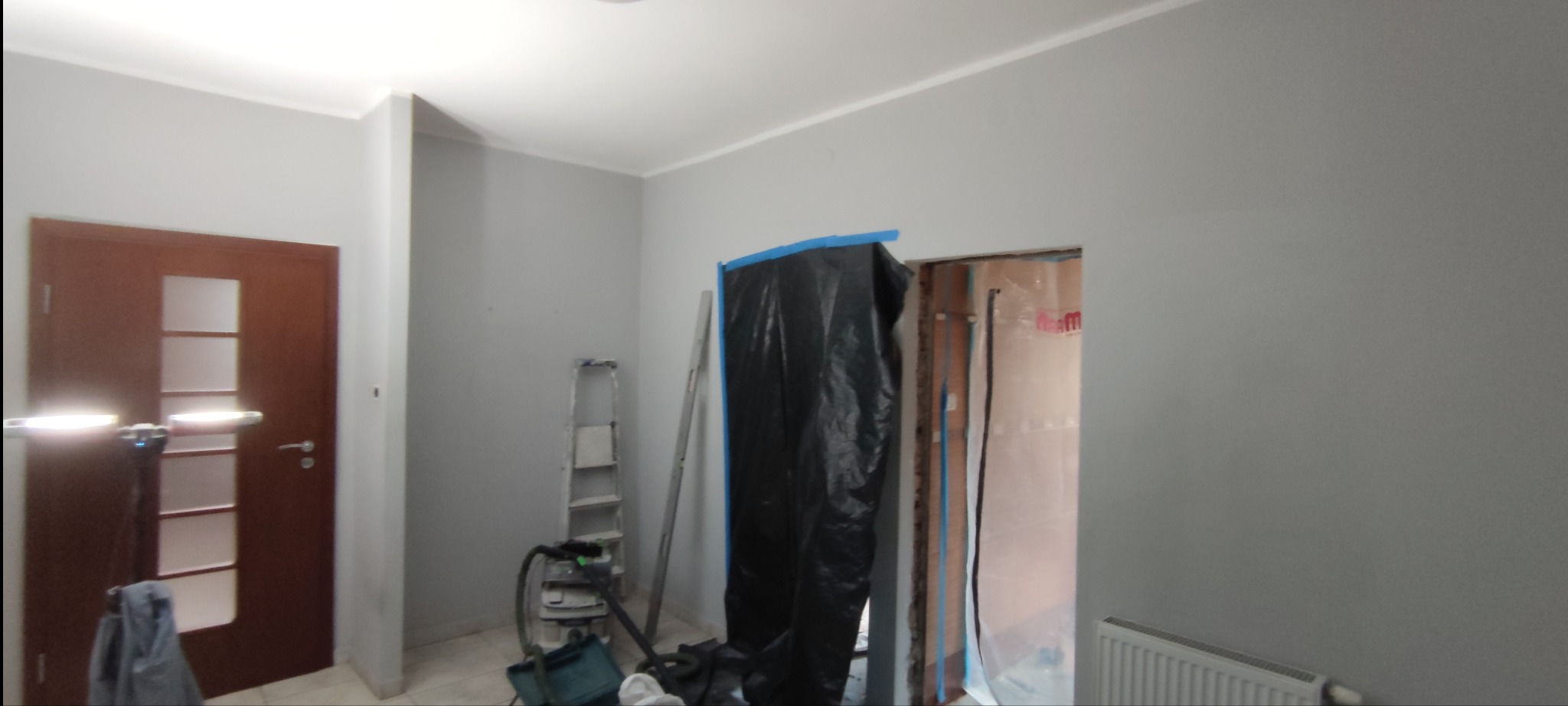 Wnętrze w trakcie odświeżania: ściany pomalowane na szaro, oklejone folią drzwi, drabina i odkurzacz na podłodze.