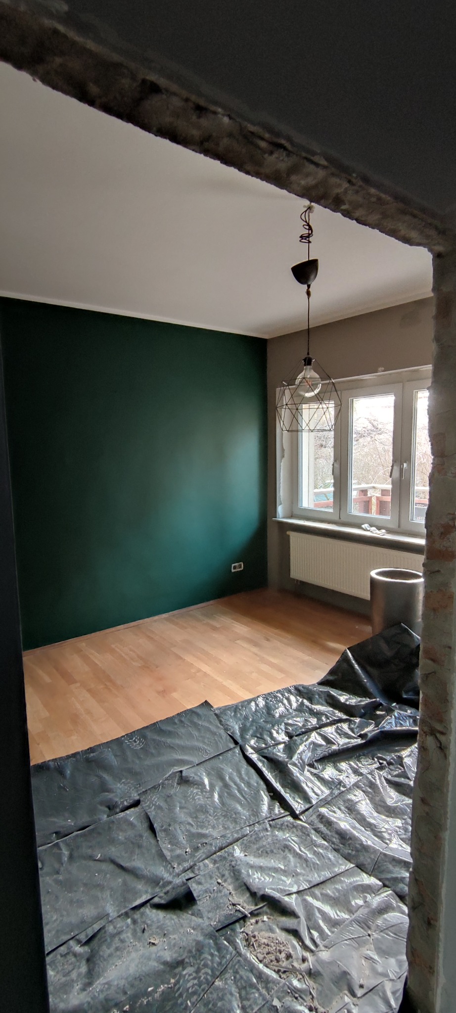 Pokój w trakcie remontu: ściana pomalowana na ciemnozielono, podłoga zabezpieczona folią ochronną, widoczna lampa wisząca i okno z grzejnikiem.